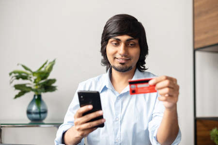 Convenience to do shopping without leaving home. Young indian man holds smartphone and credit card,...