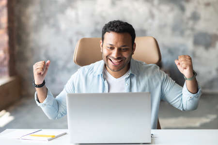 Cheerful modern young indian businessman looks at laptop screen screams happily, satisfied with the result of deal sitting at the desk in loft office space. Glad guy won game, lottery. Triumph conceptの写真素材