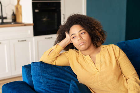 Frustrated young woman sits on the couch at home alone, upset African woman feels lonely and...