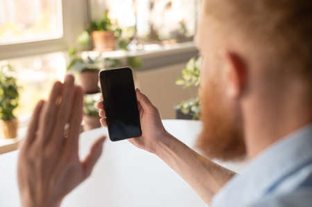 Video call concept. A man is holding mobile phone and waving hi, making video call, phone with blank...