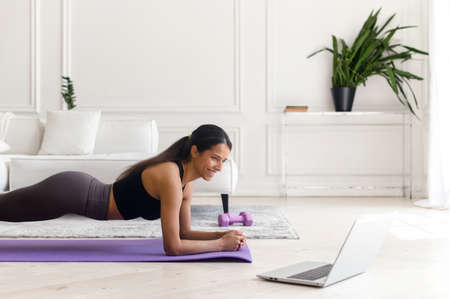 Attractive indian young woman using a laptop for watching sports video tutorial, doing plank...