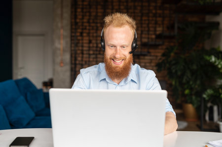 Portrait of cheerful red haired hipster guy in wireless headset using laptop computer sitting in contemporary loft style office space, man is making video call, talking online on distance, front viewの写真素材