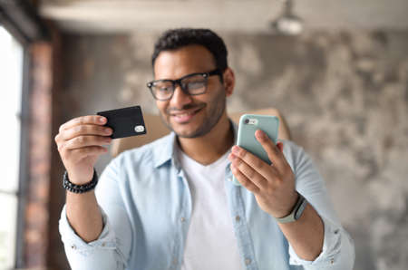 Smiling indian guy in casual wear and stylish eyeglasses holding credit card and smartphone and enjoys online shopping, making purchases online sits in the modern office. Selective focus on the cardの写真素材