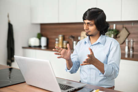 Young indian man wearing talks online, using laptop computer for video connection with colleagues or...