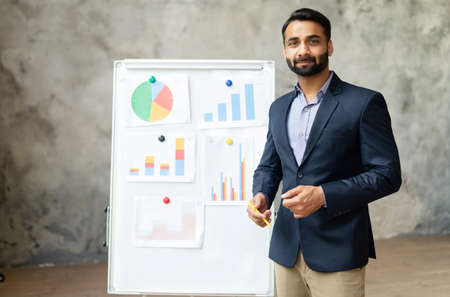 Smart and intelligent Indian man in suit stands near whiteboard with diagrams, looks at the camera,...