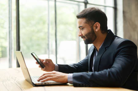 Smiling Indian male office employee in smart casual wear using smartphone during online work on the...
