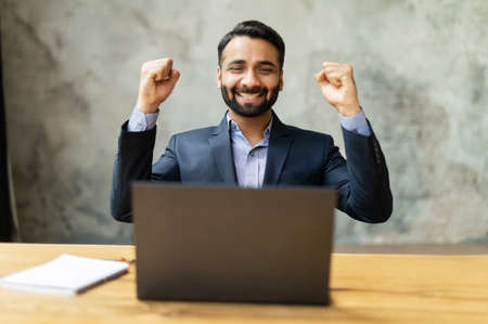 Excited and happy Indian businessman celebrating good deal or win sitting at desk, looks at the laptop screen, male entrepreneur in formal suit raising fists in triumph gesture, business going wellの写真素材