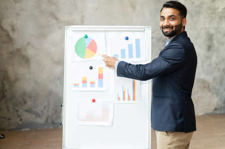 Cheerful Indian male tutor teaching students online, mixed-race man points at the whiteboard and...
