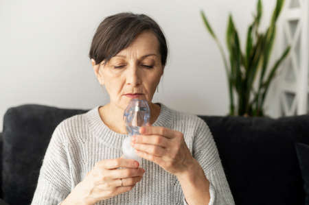 Side view a senior older woman is using inhaler for flu and cold treatment sitting on the sofa at home. Healthcare and medicine conceptの写真素材