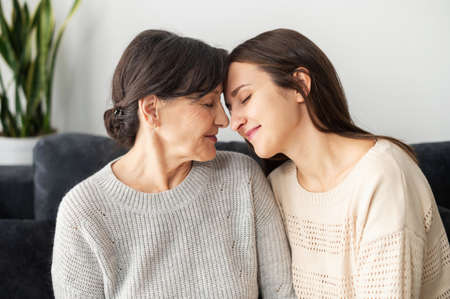 Grown daughter and senior mother are sitting on the couch in the embrace at home, hug and support...