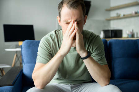 Upset young man sitting on the couch at home with hands on face, covering eyes with palms, suffers...