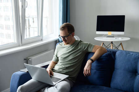 Handsome young European man with beard sitting on the sofa with laptop, at home, searching in the...