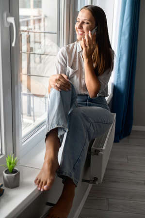 Vertical portrait of charming woman talking on the smartphone sitting on the window cill at home....