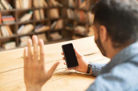 Video call concept. Indian man is holding mobile phone and waving hi, making video call, phone with blank empty screen, using new mobile app for video connection, back view, copy space, templateの写真素材
