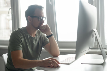 Serious focused determined young bearded businessman or freelancer in glasses and casual clothes sitting at kitchen table, using trendy PC, working on project, researching, writing email to clientの写真素材