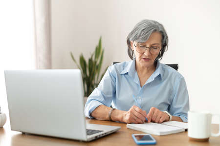 Serious mature senior female office worker business lady listening to an online webinar on laptop, taking notes in notebook, working on freelance project at home, remote work, learning at home conceptの写真素材