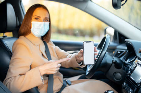 Young woman in surgical mask showing mobile phone with empty blank screen for mock up sitting on...