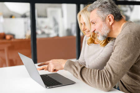 Cheerful middle-aged couple spends leisure time online with a laptop at home. Handsome grey-haired man points finger at the screen, beautiful blonde woman watching and laughs. Side viewの写真素材