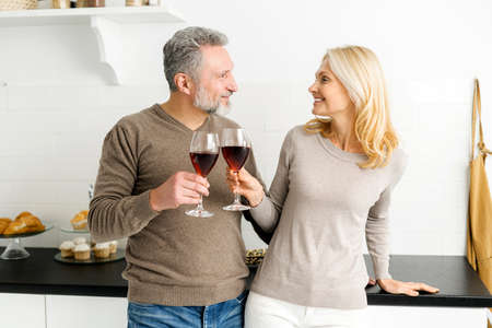 Romantic senior couple spends evening together, standing in the kitchen with glasses of red wine, handsome grey-haired man and charming mid-age woman look to each other, making cheers with glassesの写真素材