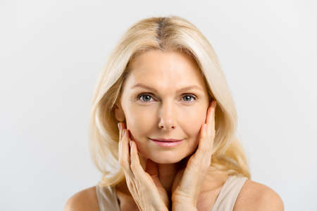 Beautiful middle-aged blonde woman with fresh pure healthy skin holding face with hand palms with natural manicure. Well-looking mature lady looks at camera, small wrinkles near eyes, anti aging careの写真素材