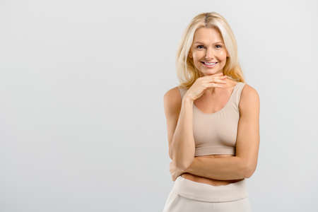 Happy carefree middle-aged lady stands in studio, isolated on grey. Charming beautiful active and fit mature blonde resting chin with hand, looks at the camera, smiles toothy. Body and face aging careの写真素材