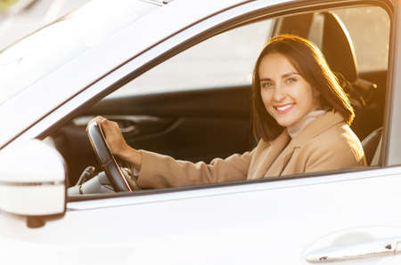 A young woman in coat is driving a car. Smiling female looks at camera and smiles in warm natural...