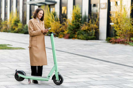 Happy woman in autumn coat using smartphone for renting modern electric scooter with an application,...