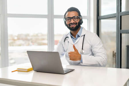 Headshot of intelligent professional Indian male doctor wearing glasses, medical white gown, headset and stethoscope, looking at the camera and shows thumb up, therapist physician in modern clinicの写真素材