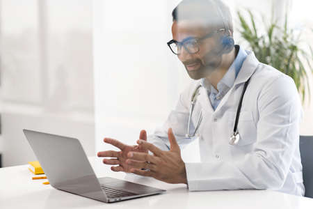 Male MD wearing white gown and stethoscope, communicating with a patient via video calling on...