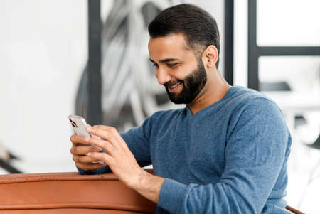 Cheerful Indian guy spends leisure scrolling news feed on the smartphone, bearded guy holds mobile...