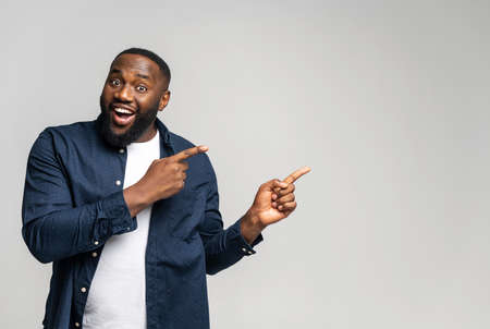 Cheerful overjoyed African-American guy points fingers at empty copy space, isolated on grey background. Multiracial man recommends deal, pays yours attention, presenting new product, advertisingの写真素材