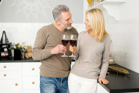 Romantic middle-aged couple holding glasses of wine look at each other and smiles standing in the kitchen at home, mature grey-haired man and charming mid-age woman celebrating anniversary, have dateの写真素材