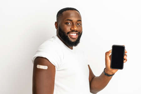 Vaccinated African-American man showing smartphone with blank screen, medical patch after injection on shoulder, black male getting vaccine dose against , qr code, vaccine certificate. Mockupの写真素材