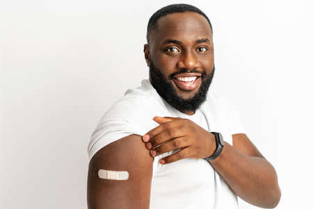 Cheerful vaccinated African-American man showing arm with medical patch and laughs, black male getting vaccine dose against , plaster on her shoulder, isolated on white. Healthcare conceptの写真素材