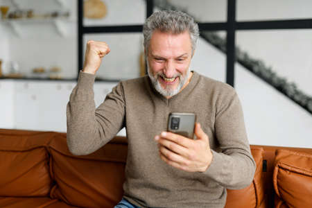 Senior grey-haired man is using smartphone for watching sports soccer match or play video game...