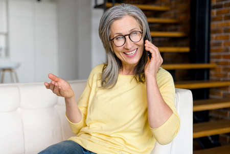 Portrait of charming senior woman talking on smartphone sitting on the sofa, friendly mature gray-haired lady listening to caller, staying in touch with her family in pandemic, has phone conversationの写真素材