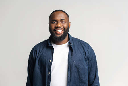 Smile. Cheerful glad African-American guy in casual blue shirt over white t-shirt looks at the camera isolated on white, happy black guy smiling with wide toothy smile. Studio shotの写真素材