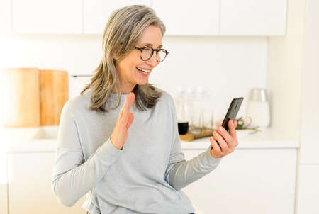 Happy smiling mature woman sitting in the kitchen, relaxing and talking to her friend or family via...