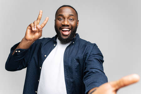 Hilarious young African-American man smiling and looking at camera, taking selfie isolated on gray background, excited happy black guy laughing at the camera, showing victory gestureの写真素材
