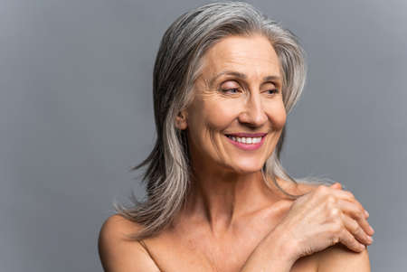Winsome senior gray-haired woman isolated on gray. Joyful beautiful mature lady posing in studio with naked shoulders, looks away and laughing, has a good mood. Well-being and healthの写真素材