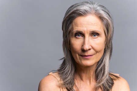 Sophisticated elderly woman with naked shoulders isolated on gray-background. Close-up portrait of well-looking mature aged lady with gray silver hair looking at camera with serious faceの写真素材