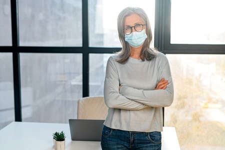 Close-up portrait of serious senior woman wearing surgical medical mask stands with arms crossed in...