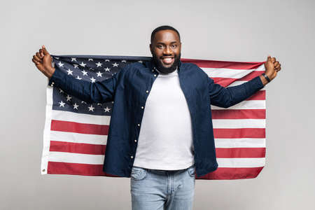 Happy African American man fighter for rights holding wide open United States flag behind back, smiling and cheerful guy protesting against racism, isolated on gray wall, black lives matter conceptの写真素材