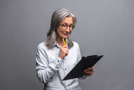 Modern elderly senior woman in formal wear taking notes standing with clipboard isolated on gray, mature female office employee writing down plans and ideas, checking up tasksの写真素材
