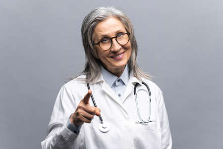 Smiling mature gray-haired female doctor in white medical gown, with stethoscope isolated on grey background points finger at you, choosing you, MD pays attention at your health and medical insuranceの写真素材