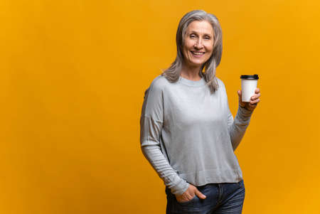 Smiling senior woman in casual wear holding paper cup of coffee isolated over yellow background, mature happy gray-haired lady standing and looking at camera, enjoying coffee to-goの写真素材