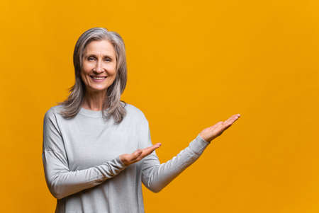 Satisfied modern mature woman presenting novelty isolated on yellow, senior gray-haired lady points aside with both hands at copy space, advertising new product, pointing with palms at blank spaceの写真素材