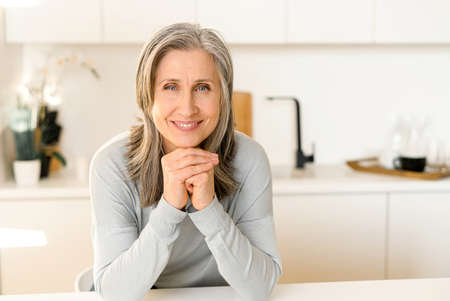 Joyful senior mature full of energy woman in casual blue pullover in the modern kitchen, gray-haired...