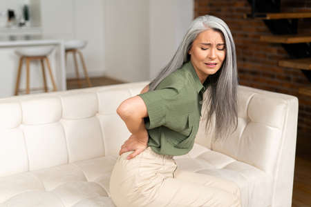 Upset mature 50s woman with gray-hair suffering from back pain sitting on sofa, middle-aged female...