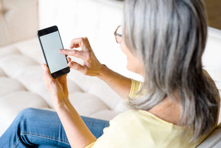 Back view a woman holding smartphone with an empty screen, senior gray-haired woman sits on couch...